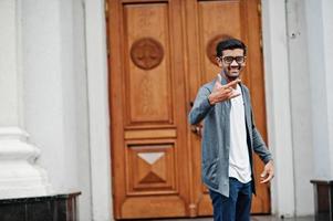elegant indisk man på glasögon bär casual poserade utomhus mot dörren till byggnaden, visa rock fingrar tecken. foto