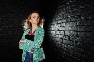 snygg blond tjej i jacka och jeans med anteckningsbok dagbok vid händerna mot tegel svart vägg på studio. foto