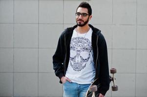 street style arabisk man i glasögon med longboard poserade mot grå vägg. foto