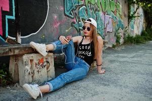 snygg casual hipster flicka i keps, solglasögon och jeans slitage, lyssnar musik från hörlurar av mobiltelefon mot stor graffiti vägg. foto