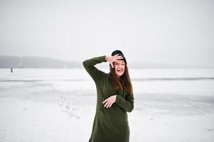 rolig flicka bär på lång grön tröja, jeans och svarta huvudbonader visar en tunga vid frusen sjö i vinterdag. foto