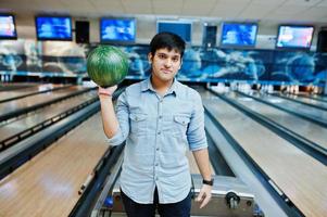 snygg asiatisk man i jeansskjorta stående vid bowlinghall med boll till hands. foto