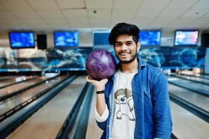 elegant skägg asiatisk man i jeansskjorta stående vid bowlinghall med boll till hands. foto