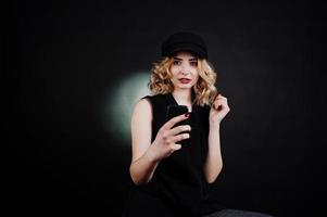 studio porträtt av blond tjej i svart slitage och mössa med mobiltelefon till hands mot mörk bakgrund. foto