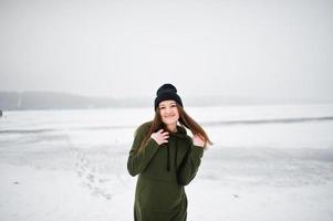 rolig flicka bär på lång grön tröja, jeans och svarta huvudbonader, vid frusen sjö i vinterdag. foto