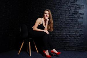 stilig brunett tjej bär på svarta och röda höga klackar, sitter och poserar på stol i studio mot mörk tegelvägg. studio modell porträtt. foto