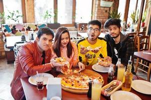 grupp asiatiska vänner som äter pizza under fest på pizzeria. glada indianer har roligt tillsammans, äter italiensk mat och sitter på soffan. foto
