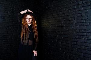 studiofotografering av flicka i svart med dreads, vid glasögon och hatt på tegelbakgrund. foto