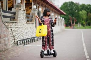 vacker afrikansk amerikansk kvinna som använder segway eller hoverboard och mobiltelefon. svart flicka med gul duk eco väskor återvinning symbol. foto