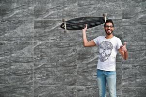 street style arabisk man i glasögon med longboard poserade mot grå vägg. tummen upp show. foto