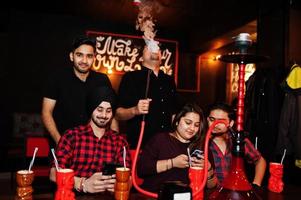 grupp indiska vänner som har kul och vila på nattklubben, dricker cocktails och röker vattenpipa, tittar på mobiltelefoner. foto