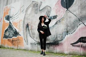 sensuell tjej helt i svarta, röda läppar och hatt. goth dramatisk kvinna håller vit krysantemum blomma mot graffiti vägg. foto