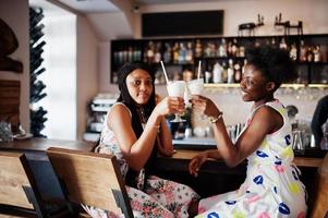 två svarta afrikanska flickvänner på sommarklänningar dricker och hejar på milkshakecocktails i baren. foto