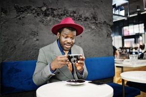 snygg afrikansk amerikansk man modell i grå jacka slips och röd hatt dricka kaffe på café. foto