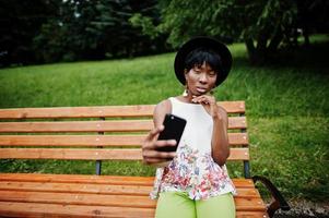 fantastisk afrikansk amerikansk modellkvinna i gröna byxor och svart hatt poserade på bänken i parken och gör selfie på mobiltelefon. foto