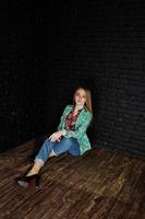 snygg blond tjej i jacka och jeans mot tegelsvart vägg på studio. foto