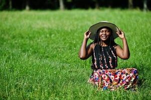 porträtt av underbar afrikansk amerikansk kvinna 20-talet i sommarhatt sitter på grönt gräs i parken. foto