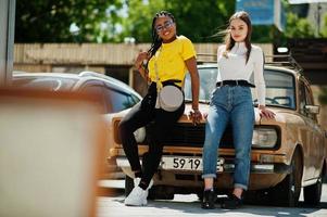 vit kaukasisk tjej och svart afroamerikan tillsammans mot gamla retrobil. världsenhet, raskärlek, förståelse i tolerans och samarbete mellan raser och mångfald. foto