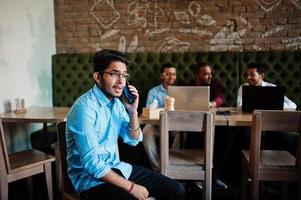 grupp av fyra sydasiatiska män poserade på affärsmöte i café. indianer arbetar med bärbara datorer tillsammans med olika prylar och pratar. indisk man med mobiltelefon. foto