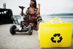 vacker afrikansk amerikansk kvinna sitter nära segway eller hoverboard. svart flicka med gul duk eco väskor återvinning symbol. foto