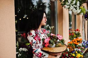 sommar porträtt av brunett flicka i rosa glasögon och hatt mot blommor butik. foto