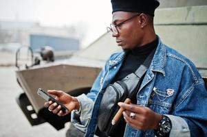 afrikansk amerikansk man i jeansjacka, basker och glasögon, rökte cigarr och poserade mot btr militärbepansrade fordon, med mobiltelefon till hands. foto