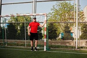 ung brutal skäggig muskulös man bär på röd skjorta, shorts och keps på stadion. foto