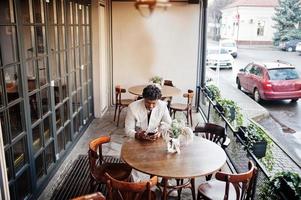 snygg afro man i beige gammal skolkostym sitter på café med mobiltelefon. fashionabla ung afrikansk man i ledig jacka på bar överkropp. foto