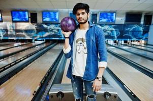 elegant skägg asiatisk man i jeansskjorta stående vid bowlinghall med boll till hands. foto