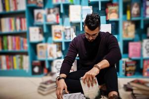 lång smart arab student man, bära på violett turtleneck och glasögon, på biblioteket sitter mot bokhyllor. foto