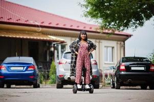 vacker afrikansk amerikansk kvinna som använder segway eller hoverboard. svart flicka på dubbla hjul självbalanserande elektrisk skoter mot bilparkering. foto