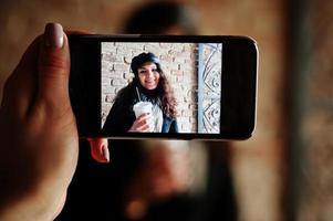 lockig mexikansk tjej i lädermössa och plastkopp kaffe till hands på skärmen på mobiltelefonen. foto