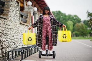 vacker afrikansk amerikansk kvinna som använder segway eller hoverboard. svart flicka med gul duk eco väskor återvinning symbol. foto