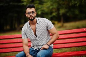 fashionabla långa arabiska skägg man bär på skjorta, jeans och solglasögon sitter på röd bänk i parken. foto
