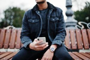 stilig och fashionabel indisk man i svart jeansjacka poserade utomhus, sittande på bänken med mobiltelefonen till hands. foto