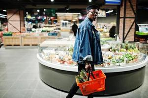 snygg casual afrikansk amerikansk man på jeansjacka och svart basker håller korg med ananas i frukter ekologisk del av stormarknaden. foto