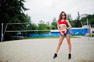 sexig lockig modell tjej i röd topp, jeans jeansshorts, kopp och solglasögon poserade på beachvolleyplan. foto