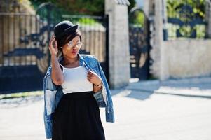 snygg afrikansk amerikansk modell i glasögonhatt, jeansjacka och svart kjol poserade utomhus. foto