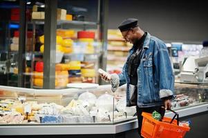 snygg casual afroamerikansk man på jeansjacka och svart basker håller korg, står nära ostkyl och shoppar i snabbköpet. foto