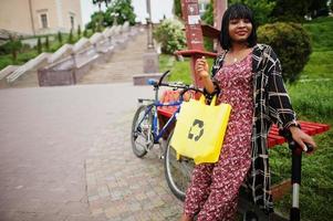 vacker afrikansk amerikansk kvinna som använder segway eller hoverboard. svart flicka med gul duk eco väskor återvinning symbol. foto