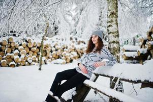 söt lockig tjej i tröja och huvudbonader sitter på bänken på snöig skogspark på vintern. foto