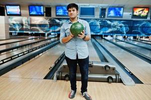 snygg asiatisk man i jeansskjorta stående vid bowlinghall med boll till hands. foto