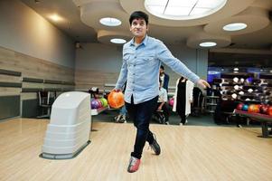 sydasiatisk man i jeansskjorta står vid bowlinghall med boll på händerna. killen förbereder sig för ett kast. foto
