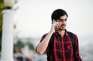 ung indisk student man på rutig skjorta och jeans med ryggsäck talar i mobiltelefon. foto