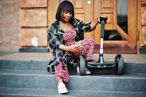 vacker afrikansk amerikansk kvinna sitter nära segway eller hoverboard. svart tjej med dubbla hjul självbalanserande elektrisk skoter med mobiltelefon. foto