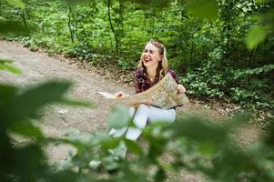 porträtt av en positiv ung vacker blondin som sitter på marken med en karta i händerna i skogen. foto