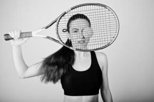 svart och vitt porträtt av vacker ung kvinna spelare i sportkläder håller tennisracket medan stående mot vit bakgrund. foto