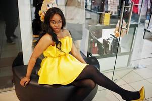snygg afrikansk amerikansk kvinna på gul klänning sitter på en puff mot montrar på köpcentret. foto