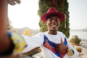 snygg trendig afro frankrike man i röd hatt och vit outfit poserade på höstdagen. svart afrikansk modell kille hålla kameran och göra selfie. foto