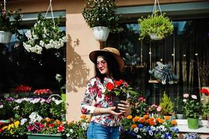 sommar porträtt av brunett flicka i rosa glasögon och hatt mot blommor butik. foto
